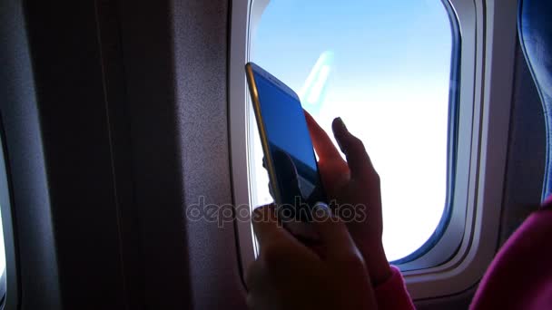 Vista desde la ventana del avión, se puede ver la silueta oscura de las manos femeninas que trabajan en un gadget, smartphone, teléfono móvil, movimiento de los dedos. Cielo visible, nubes, clima claro, día soleado — Vídeos de Stock