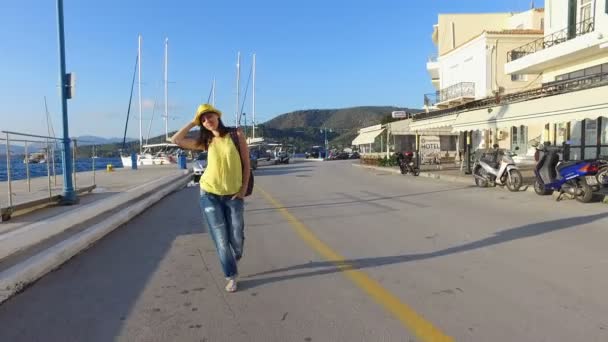 Chica turista morena en una camiseta amarilla, vaqueros azules, un sombrero amarillo y con una mochila de colores camina a lo largo del paseo marítimo, al atardecer, a la luz del sol, la luminosidad, en verano, la isla de Poros, Grecia. Lo es. — Vídeos de Stock