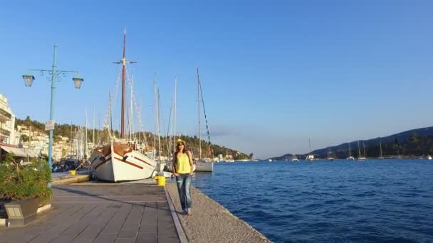 Turista bonita, jovem morena em óculos de sol em uma camiseta amarela, jeans azul, um chapéu amarelo e com uma mochila colorida caminha ao longo da orla, ao pôr do sol, à luz do sol, radiância, no — Vídeo de Stock