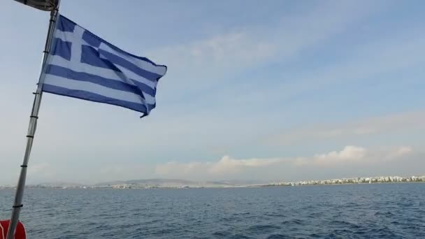 Di latar belakang lanskap pegunungan dan Laut Tengah, bendera biru dan putih Yunani bergoyang-goyang dalam angin, di atas kapal pesiar, di kejauhan terlihat pantai Athena — Stok Video