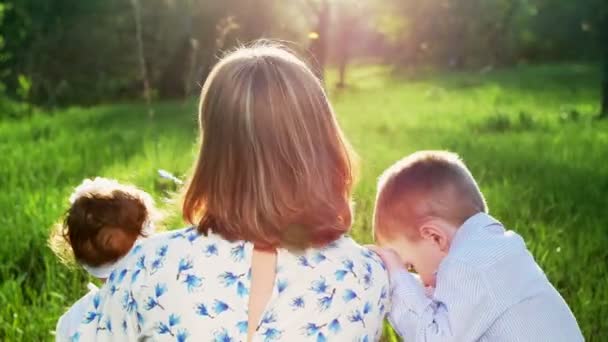 W promieniach słońca, pod światło, o zachodzie słońca, latem w parku, młoda matka, blond kobieta wieje bańki mydlane dla jej dzieci, jej dwóch synów. Chłopcy są zabawne, ciekawe — Wideo stockowe