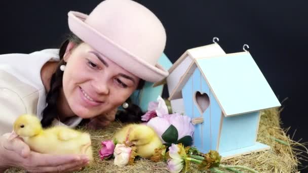 Retrato, una hermosa joven con dos coletas y un divertido sombrero rosa jugando con pequeños patitos amarillos. En el fondo un pajar, casas de pájaros de colores, y flores . — Vídeo de stock