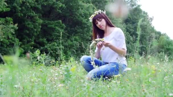Verão, em meio a um gramado de camomila, em uma floresta, uma jovem mulher, uma morena tece uma coroa de camomila — Vídeo de Stock