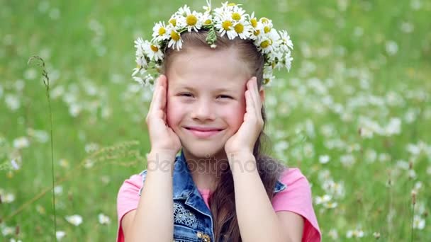 Sur une pelouse de camomille, une fille douce dans une couronne de marguerites, souriante, pressant ses mains sur ses joues — Video