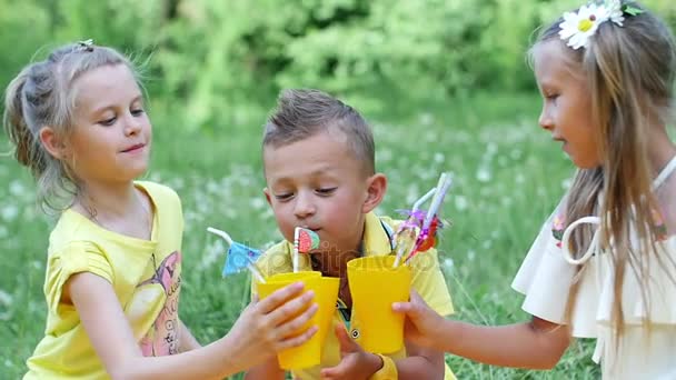 En un prado de manzanilla, cerca de un bosque, en la hierba, hay tres niños en un yeso amarillo, que beben bebidas dulces a través de túbulos. Tienen un picnic, se divierten, son engañados , — Vídeos de Stock