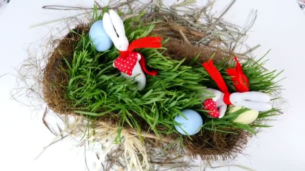 Крупный план, вид сверху, ротация, праздничная пасхальная композиция состоит из цветных яиц, зеленой молодой травы и мягких фигурок белых пасхальных кроликов в красных шарфах — стоковое видео