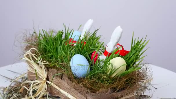 Su sfondo bianco, rotazione, La composizione pasquale festiva è composta da uova colorate, erba giovane verde e morbide figure di coniglietti pasquali bianchi in sciarpe rosse — Video Stock