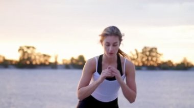 Güzel, ince genç sarışın bir kadın fitness konusunda nişanlı atlar, güç egzersizleri gerçekleştirir. Şafakta, kumlu taş ocağı plajda güneş ışığı, güneş karşı çekim video.