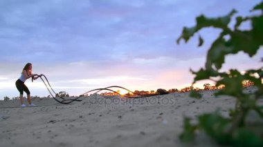 Genç, atletik, sarışın kadın, güç egzersizleri kalın, sportif bir ip yardımı ile gerçekleştirir. Şafakta, kumlu taş ocağı plajda güneş ışığı, güneş karşı çekim video.