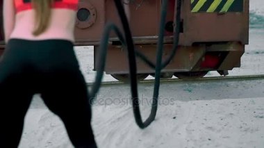 atletik kadın, güç egzersizleri kalın, sportif bir ip yardımı ile gerçekleştirir. Bir ip close-up. Şafakta, kumlu taş ocağı plajda güneş ışığı altında yakınındaki bir kargo vinç,