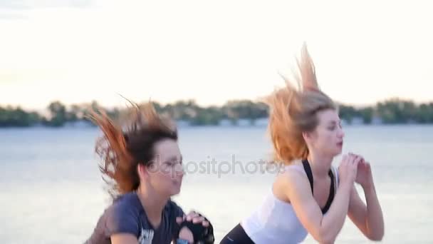 As mulheres jovens belas, esbeltas envolvem-se na aptidão, pulam, executam exercícios de força. Ao amanhecer, na praia da pedreira , — Vídeo de Stock