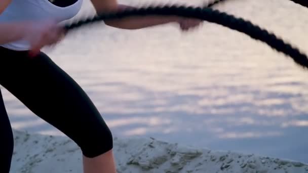 Jeune, athlétique, femme blonde, effectue des exercices de force à l'aide d'une corde épaisse et sportive. À l'aube, à la plage de la carrière de sable, à la lumière du soleil, tournage vidéo contre le soleil . — Video