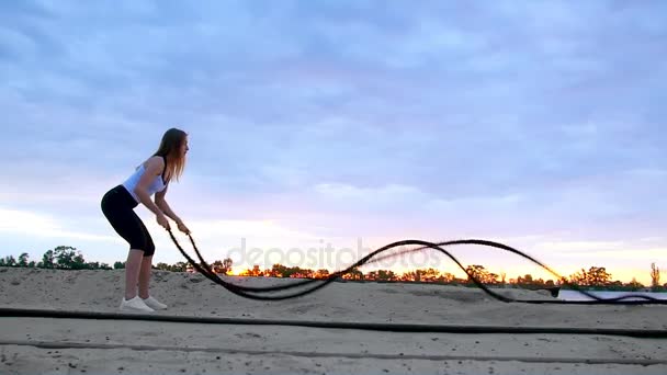 Jovem, atlética, loira, realiza exercícios de força com a ajuda de uma corda espessa e esportiva. Ao amanhecer, na praia de areia pedreira, à luz do sol, filmagem de vídeo contra o sol . — Vídeo de Stock