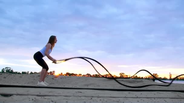 Mujer joven, atlética, rubia, realiza ejercicios de fuerza con la ayuda de una cuerda gruesa y deportiva. Al amanecer, en la playa de la cantera de arena, a la luz del sol, video rodaje contra el sol . — Vídeo de stock