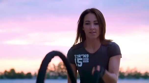 Jong, sportief, vrouw, voert kracht oefeningen met behulp van een dikke, sportieve touw. Bij dageraad, op het zandstrand steengroeve strand, in het zonlicht, video schieten tegen de zon. — Stockvideo