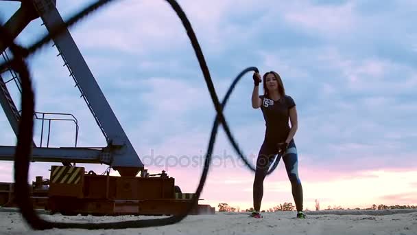 Jong, sportief, vrouw, voert kracht oefeningen met behulp van een dikke, sportieve touw. Bij dageraad, op het zandstrand steengroeve strand, in het zonlicht, in de buurt van een lading kraan, — Stockvideo