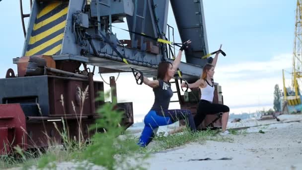 На світанку, біля вантажного крана, дві молоді красиві, спортивні дівчата займаються вправами з ременями підвіски trx .Fitness вправи для відпрацювання м'язів всього тіла . — стокове відео