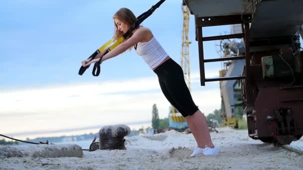 Hermosa mujer atlética haciendo ejercicio con sistema trx. Ella hace flexiones de brazos de entrenamiento con correas de fitness trx. Al amanecer, cerca de una grúa de carga, en la playa de la cantera , — Vídeos de Stock