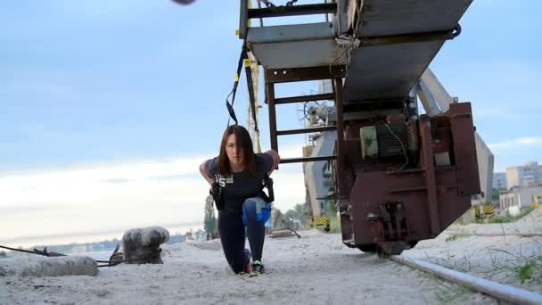 Hermosa dama ejercitando sus músculos con la ayuda de correas de suspensión TRX. Al amanecer, cerca de una grúa de carga, en la playa de la cantera , — Vídeos de Stock