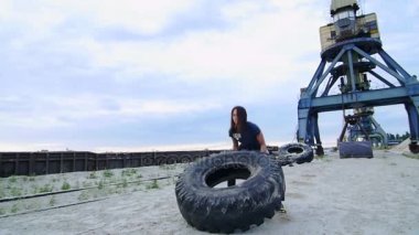 Atletik bir kadın büyük ağır traktör tekerleğini kullanarak egzersizleri gerçekleştirir, kasları koçlar. Atıyor. Arka plan üzerinde olabilir görmedim kargo vinç, kargo liman, şafak.