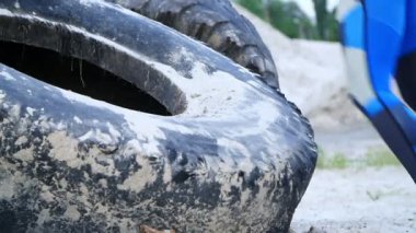 Yakın çekim, t-shirt atletik kadın, mavi leggens büyük bir traktör tekerleği kullanarak güç egzersizleri gerçekleştirir, sahilde atlama, kargo limanı, kum üzerinde. şafak vakti. Yavaş çekim.