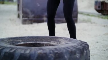 athletic woman in a grey top and black leggings performs strength exercises using a large heavy tractor wheel, trains her muscles. On the beach, cargo port, on the sand