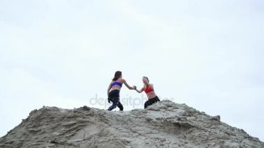 İki genç, atletik kadın açık havada egzersiz yapıyor, taş ocağının kumlu tepesinde. Sahilde, kargo limanında, kumda.