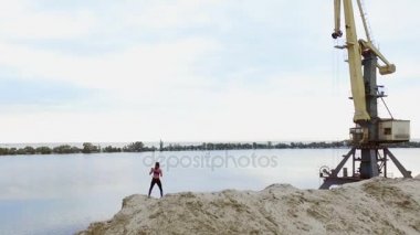 Genç, atletik kadın boks, dışarıda, taş ocağının kumlu tepesinde egzersiz yapıyor. Arka planda nehir, kargo vinci, şafak görülebilir. Aero video çekimi