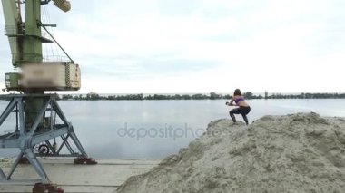 Genç, atletik bir kadın spor yapıyor Squats, taş ocağının kumlu tepesinde, sahilde. Arka planda nehir, kargo vinci, şafak görülebilir. Aero video çekimi