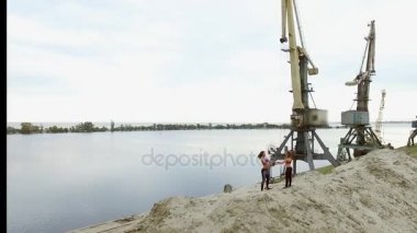 İki genç, atletik kadın, sahilde, taş ocağının kumlu tepesinde egzersizler yapıyor. Arka planda nehir, kargo vinçler, şafak görülebilir. Aero video çekimi.