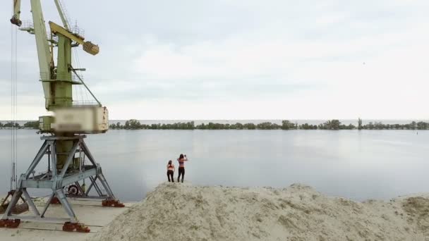 Две молодые атлетичные женщины делают упражнения на растяжку, разминку, на песчаной вершине карьера, на пляже. На заднем плане видны река, грузовые краны, рассвет. Аэровидеосъемка . — стоковое видео