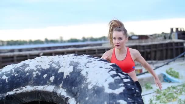 Eine junge athletische Frau macht Übungen mit einem großen schweren Traktorrad, trainiert ihre Muskeln. sie springt. Im Hintergrund sieht man Frachtkräne, Frachthafen, Morgendämmerung. — Stockvideo