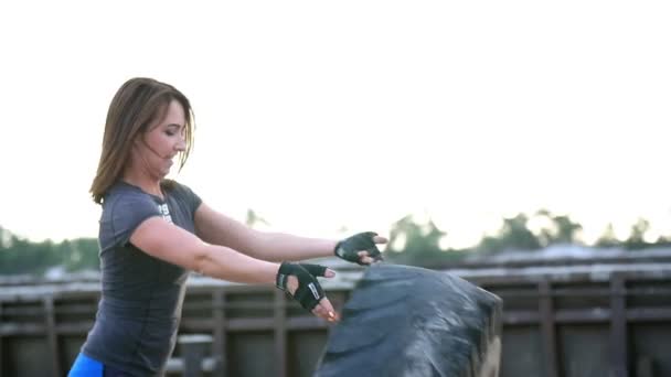 En ung kvinna som atletisk utför övningar med en stor tung traktor hjul, coachar sina muskler. Kastar den. I bakgrunden kan vara sett styckegodskranar, cargo port, dawn. — Stockvideo