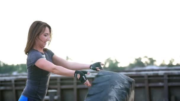 Une jeune femme sportive effectue des exercices à l'aide d'une grande roue de tracteur lourd, entraîne ses muscles. Jette-le. Sur le fond peut être vu des grues à cargaison, port de fret, aube . — Video