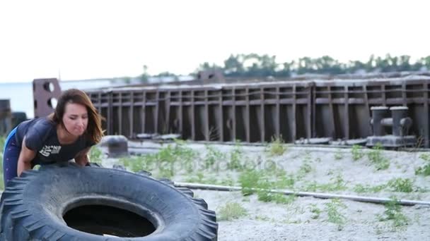 Une jeune femme sportive effectue des exercices à l'aide d'une grande roue de tracteur lourd, entraîne ses muscles. Jette-le. Sur le fond peut être vu des grues à cargaison, port de fret, aube . — Video