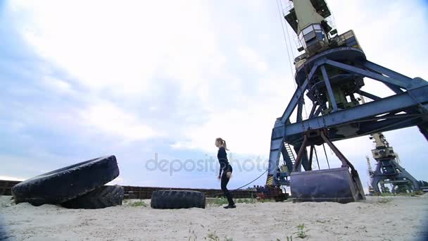 Donna atletica in un top grigio e leggings neri esegue diversi esercizi di forza con una grande ruota trattore pesante, allena i muscoli. dietro è gru di carico, porto di carico, alba — Video Stock