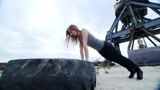 Mujer atlética en una parte superior gris y polainas negras realiza ejercicios de fuerza utilizando una rueda tractora grande, haciendo flexiones brazos de entrenamiento. En la playa, puerto de carga, en la arena — Vídeos de Stock