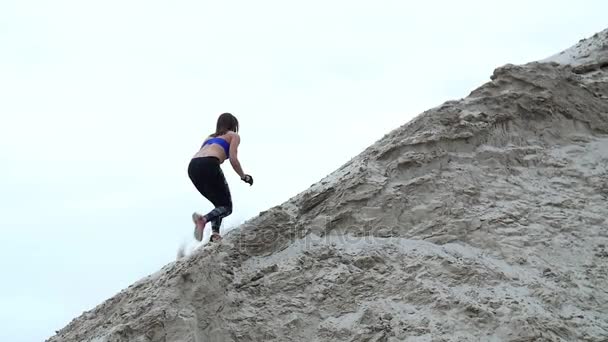 Genç bayan sporcu kumlu bir dağa tırmanır. Arka planda kargo vinçleri, kargo iskelesi, şafak görülebilir.. — Stok video