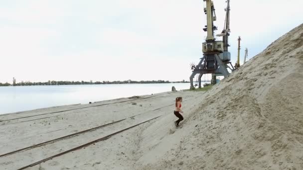 Young woman athlete climbs a sandy mountain. On the background can be seen the river, cargo cranes, cargo port, dawn. — Stock Video