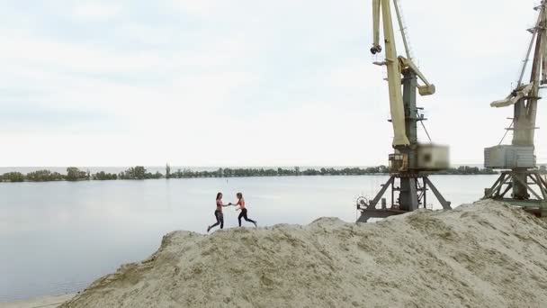 Dos mujeres jóvenes y atléticas haciendo ejercicios al aire libre, en la parte superior arenosa de la cantera. En el fondo se puede ver el río, grúas de carga, amanecer. Aero rodaje de vídeo . — Vídeo de stock