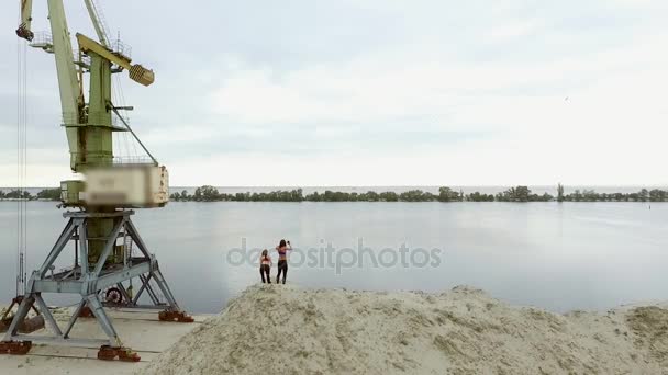 Dos mujeres jóvenes y atléticas haciendo ejercicios de estiramiento, calentamiento, en la parte superior arenosa de la cantera, en la playa. En el fondo se puede ver el río, grúas de carga, amanecer. Aero rodaje de vídeo . — Vídeo de stock