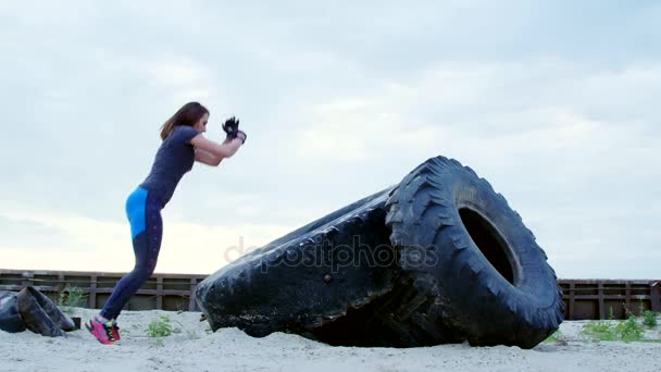 Mujer atlética en camiseta, piernas azules realiza ejercicios de fuerza utilizando una rueda tractor grande, Ella está saltando. En la playa, puerto de carga, en la arena. dawn.Slow motion — Vídeos de Stock