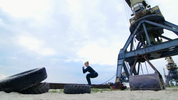 Atletisk kvinna i en svart sport kostym utför styrkeövningar med hjälp av en stor traktorhjul, tränar hennes muskler. Hon hoppar. På stranden, Last hamn, på sanden — Stockvideo