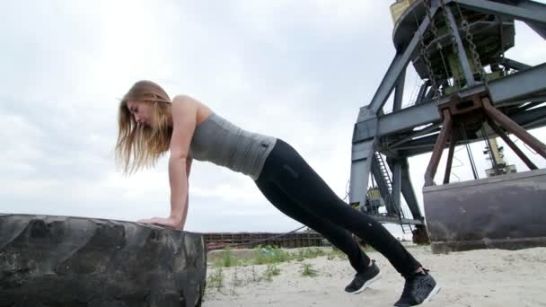 Mujer atlética en una parte superior gris y polainas negras realiza ejercicios de fuerza utilizando una rueda tractora grande, haciendo flexiones brazos de entrenamiento. En la playa, puerto de carga, en la arena — Vídeos de Stock