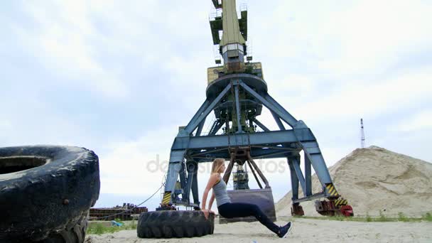 Атлетичная женщина в сером топе и черных леггинсах выполняет силовые упражнения, используя большое колесо трактора, делая отжимания тренируя руки. На пляже, в грузовом порту, на песке — стоковое видео
