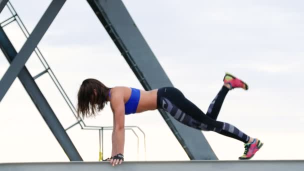 Femme sportive dans un haut bleu et leggens rayés effectue des exercices de musculation. Elle se tient sur la poutre métallique d'une grue de fret. forme physique à l'aube, sur la plage dans un port de marchandises . — Video