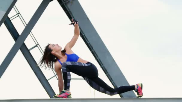 Mavi bir üst ve çizgili leggens atletik kadın gücü çalışmaları gerçekleştirir. O bir yük vinç metal kiriş üzerinde duruyor. bir kargo liman plajında şafak, Fitness. — Stok video