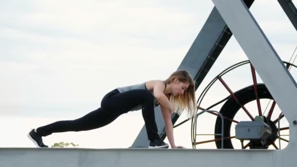 Mulher atlética em um top cinza e leggings pretos executa exercícios de força. Ela está no feixe de metal de um guindaste de frete. fitness ao amanhecer, na praia em um porto de carga . — Vídeo de Stock