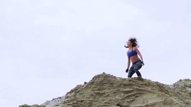 Junge, athletische Frau in blauem Oberteil und gestreiften Beinen bei Übungen, Kniebeugen, auf der sandigen Spitze des Steinbruchs, Fitness bei Sonnenaufgang, am Strand im Frachthafen. — Stockvideo