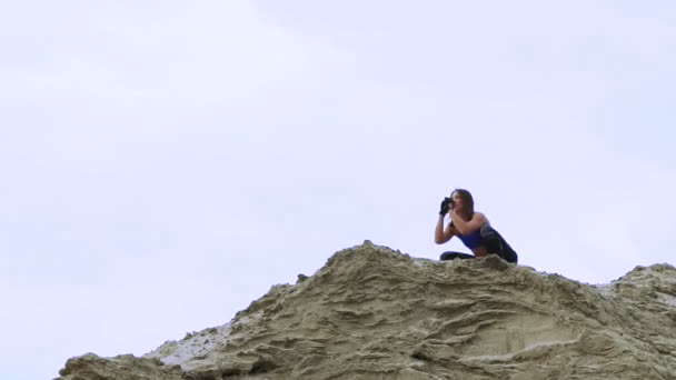 Joven, atlética mujer en un top azul y piernas rayadas haciendo ejercicios, sentadillas, en la parte superior arenosa de la cantera, fitness Al amanecer, en la playa en un puerto de carga . — Vídeo de stock